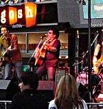 Band performing at a shopfront. Male at left is playing a guitar and is at a microphone. A fifty-something male in middle is shown partly in left profile. He has dark hair, wears sunglasses and is playing a guitar while at a microphone. A drum kit is to his left with a third band member obscured by frame cut-off. Two audience members are seen in the foreground.