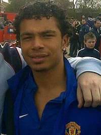 A photograph of a mixed-race man with short, curly, black hair wearing a blue shirt.