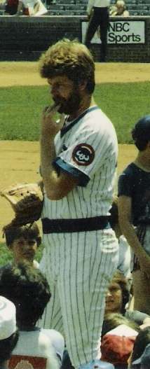 White man with beard and mustache standing on a baseball field with his hand in his mouth, wearing a white Cubs uniform with blue pinstripes and no cap.