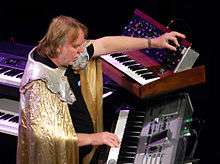 Rick Wakeman, surrounded by several keyboard instruments and wearing his customary robe, plays a keyboard with one hand and programs a Minimoog synthesizer with the other