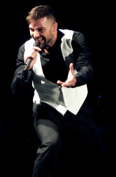 A blonde man is smiling while performing. He is holding a microphone in his right hand.