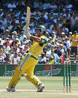 A man in a predominately yellow cricket with: a helmet, gloves, pads and a bat. He is swinging the bat as the crowd watches in the background.