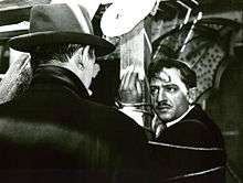 A screen capture from the film, showing a distraught César tied up to a pole staring at Tony le Stéphanois, who has his back to the camera.