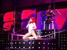 A woman with red hair wearing a white PVC body suit is performing on a raise platform. She is singing into a microphone and is handcuffed to the stage.