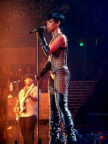 A female with medium skin and short black hair. The female is holding onto a microphone and a microphone stand with both her hands and is wearing black gloves that cover both her arms from above her elbow down. She is wearing black thigh high boots.