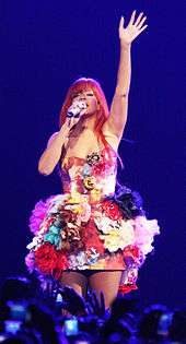 Rihanna with red, long hair and a dress with floral patterns. Her left hand is raised and her right hand holds a microphone near her mouth.