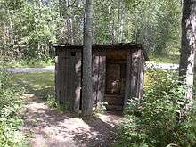 Nestled in the woods at the edge of the cleared area, the privy or outhouse has two stalls and is made of weathered wooden planks.