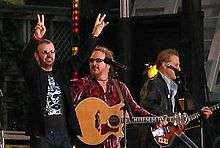Starr is on stage with two guitarists and two microphones. He is wearing sunglasses and a black and silver T-shirt, and both of his arms are raised. His right arm forms a V-shaped peace symbol.