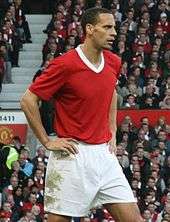 A mixed-race man with very short dark hair standing with his hands on his hips. He is wearing a red football shirt with a white V-neck and white shorts.