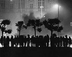 A line of people silhouetted against a building, with a plume of smoke rising behind the people.