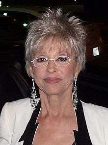 A woman is wearing glasses, a white dress, and earing is facing the camera.