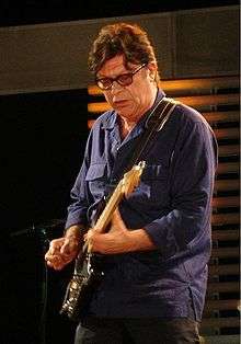 "A colour photo of Robbie Robertson on a stage. He is wearing a purple shirt while playing a six string fender guitar."