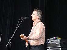 Forster is shown in upper half, left profile. He is standing, singing into a microphone and playing an electric guitar. Some musical equipment is visible behind him. His shirt is light coloured and he wears dark pants.