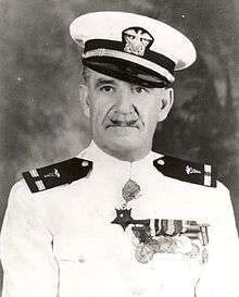Head and shoulders of a white man with a thin, upturned mustache wearing a white peaked cap with black visor and a white jacket with dark shoulder boards, a row of medals on the left breast, and a star–shaped medal hanging from the neck.
