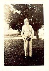 A black and white image (c.1900s) of an elderly man with white hair and a long beard dressed in a long-sleeved and suspenders standing in front of a tree.