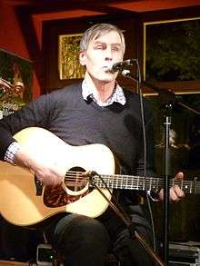 Three-quarter shot of a 55-year-old man sitting on a chair, he is singing into a microphone and playing his guitar. The guitar also has a microphone. He wears dark clothes and has short hair.