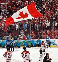 A helmetless ice hockey goaltender carrying a large Canadian flag by its pole over his head as teammates, spectators and media look on. He is wearing a white and red jersey with white pads.