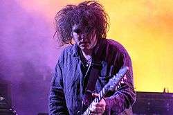 Head and shoulders shot of man, with wild, tangled hair and lipstick on, playing in a stage spotlight.