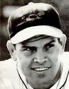 A headshot of a smiling man in a dark baseball cap with a bird on the front.
