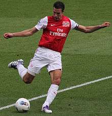 Robin van Persie, wearing a red and white football jersey and white shorts, prepares to kick a football with his right foot with both arms outstretched.