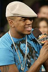 Ne-Yo wearing a beige cheese-cutter hat, blue T-shirt and a necklace, speaking into a microphone.