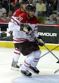 Hockey player in white and red uniform. He jumps slightly above the ice, holding his stick.