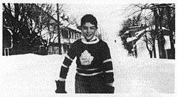 A young boy stands on a snow-covered street. He is wearing a dark-coloured sweater with a stylized maple leaf logo on the chest.