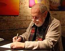 A gray-haired man seated at a table, wearing eyeglasses, a sweater, and a scarf draws with a pen in his right hand.