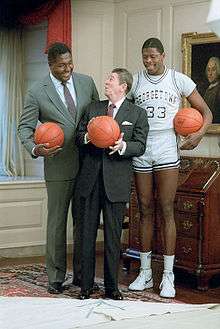 Two tall African-American men, one in a suit, one in a gray basketball uniform, stand behind a shorter elderly white male in an ornate room, with each man holding a basketball.