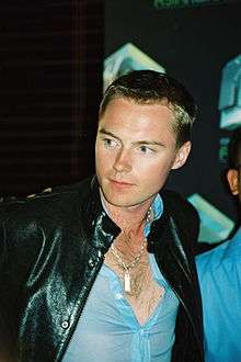 A blonde man wearing a blue T-shirt and black leather jacket.