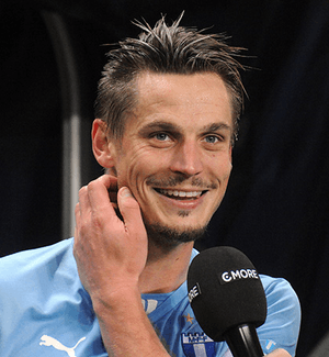 A zoomed in photograph of a man with sprawling hair and stubble, dressed in a sky blue football shirt. He is smiling brightly and itching his right cheek. He is in the process of being interviewed by a man outside the frame who is extending a black microphone.