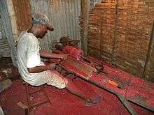 A man sits on a stool at a bench while using simple tools to spin a piece of rosewood while he carves it into a vase. Bright red sawdust covers the floor of this small rosewood vase factory.