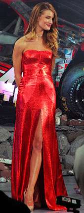 A blonde woman wearing a red dress and a gold necklace. Stones and a car can be seen behind her.