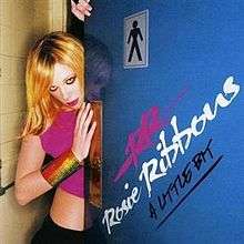 A blond woman standing behind a restroom door. She is wearing a pink top, black pant and bangles on her right hand. On the door the words "RR", "Rosie Ribbons" and "A Little Bit" are written with paint.
