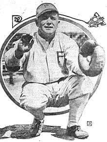 A man in a white baseball uniform with a "P" on the chest and cap is crouched waiting to catch a ball in his catcher's mitt.