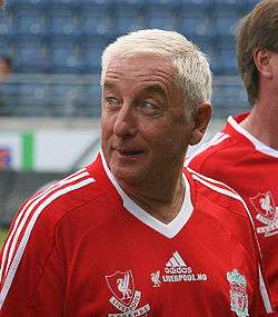 The head and upper torso of a gentleman in his late 50s. He has short white hair and is wearing a red football shirt, which has the Liverpool F.C. crest on the left breast, and a crest on the right breast that says "Liverpool Legends". A white logo of the Adidas sponsor is visible in the centre of the shirt, and three white stripes are present on the shoulder.