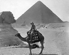 Man in military uniform and pith helmet sitting on a camel in front of the Sphinx and Pyramid