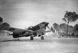 Single-engined fighter plane on airfield with propeller spinning and man sitting on left wing