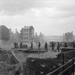 A file of soldiers walking through a blasted cityscape; only a few buildings are standing