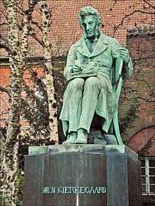 A statue. The figure is depicted as sitting and writing, with a book on his lap open. Trees and red tiled roof is in background. The statue itself is mostly green, with streaks of grey showing wear and tear. The statue's base is grey and reads "SØREN KIERKEGAARD"