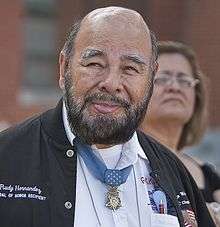 Head and shoulders of a bald man with dark beard, with a medal hanging from a blue ribbon around his neck.
