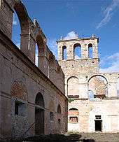 Photograph taken amid the ruins of a stone building, showing dirt instead of flooring, and rough stone walls rising three stories high with missing windows and frames. The ruins have no roof and no intermediate floors of upper levels.