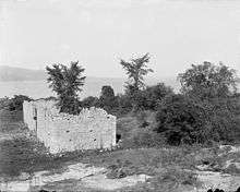 A black-and-white photograph.  Ruined wall sections are visible in the center, with a tree growing out of the center of one of the structures.  The lake is visible in the background, and hazy land is visible even further back.