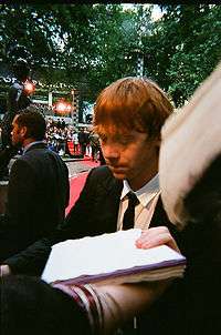 A picture of a man with red hair looking downward. A person is extending their arm to whist holding sheets of paper. Trees and red heights can be seen in the background.