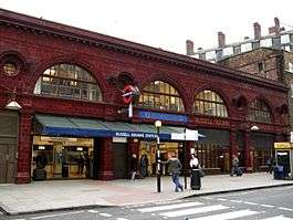 A red glazed terracotta building. The first storey above ground features four wide, storey-height semi-circular windows with smaller circular windows between above which is a dentil cornice. Below the two right-most windows, the station name, "Russell Square Station", is displayed in gold lettering moulded into the terracotta panels. A blue tiled panel above the entrance says "Underground".