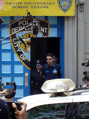 Two police officers escorting a man wearing sunglasses and a blue shirt out of a door in a wall on a city street. The wall and door are painted with the logo of the New York City Police Department. Above it is a promotional banner with the words "The NYPD and community working together". In the foreground is a police car. People taking photographs and microphones on booms intrude on the edges of the image.