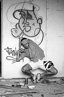 A black and white photo of a Russian popper posing mid-dance in front of a graffiti painted wall on the side of an industrial building.