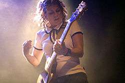 A Caucasian female with shoulder-length brown hair wearing a white shirt and dark pants strums a yellow electric guitar.