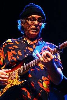 A man in a printed shirt wearing eyeglasses and a cap on his head, playing a guitar