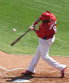 A man in a red baseball jersey, white pinstriped pants, and a red batting helmet swings at a baseball. He is standing in the batter's box on the right-hand side of home plate, and his bat is blurred in motion.
